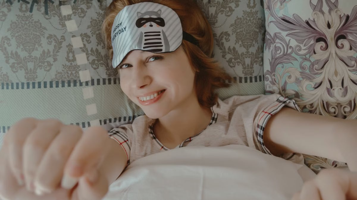 Top view of smiling female reaching out hands to camera while lying on bed under blanket and looking at camera
